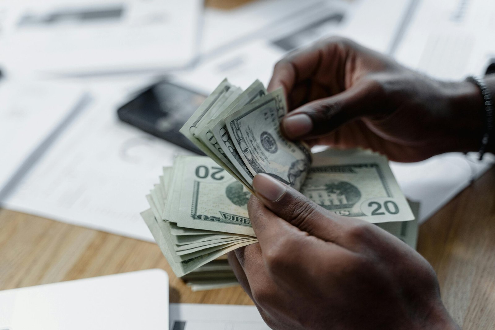 A close-up of hands counting US dollar bills on a wooden table with documents and a smartphone nearby.